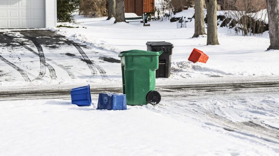 Empty wheeled garbage barrels and recycling containers are scattered/strewn near two opposite driveways along a suburban residential neighborhood street. Winter snow is on the ground and multiple slushy, messy tire tracks cris cross the driveways and the street. Must have been garbage/trash pickup day today.