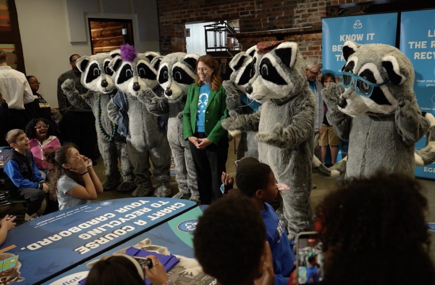 Five raccoon mascots and a woman interact with excited children at a recycling event