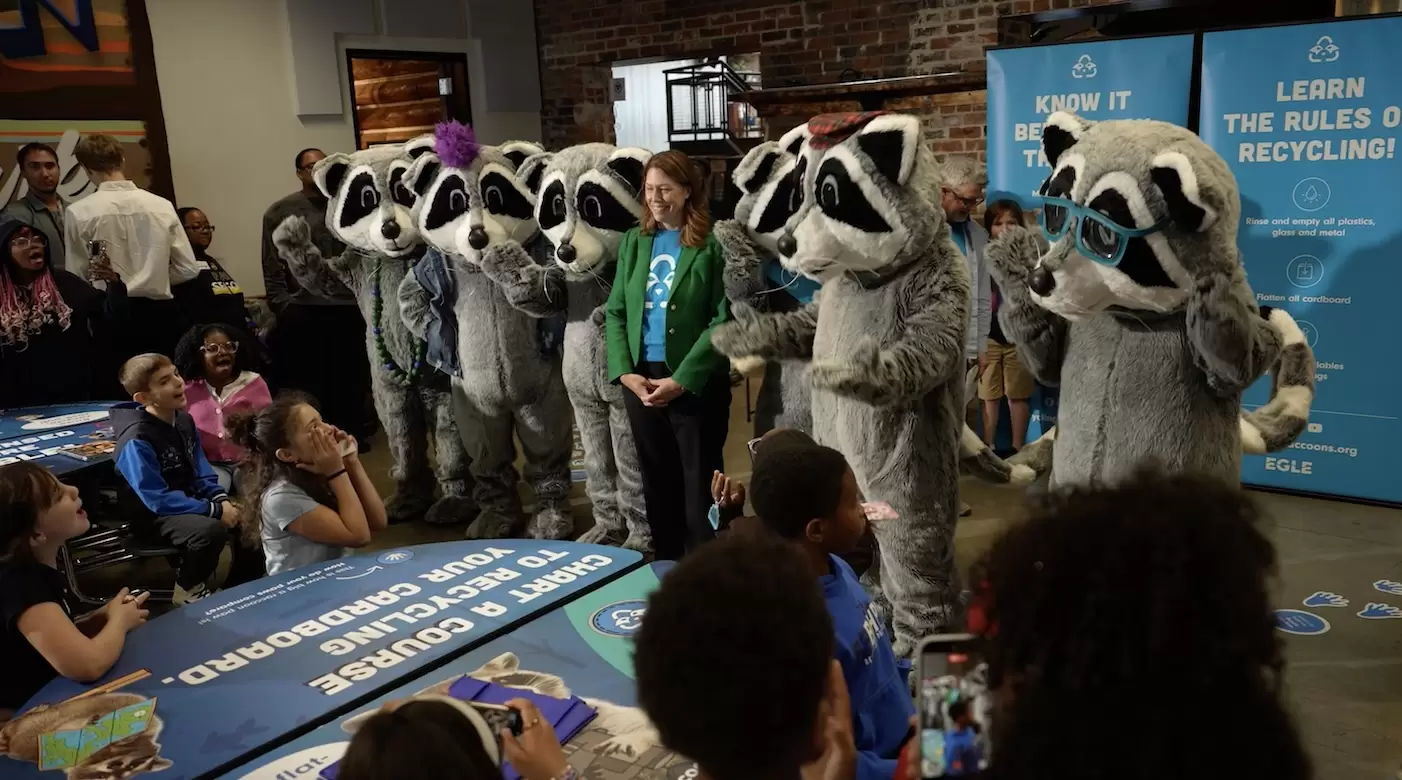 Five raccoon mascots and a woman interact with excited children at a recycling event